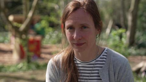 A mid shot of Victoria Bassett. She has long brown hair and is looking into the camera. She is wearing a grey cardigan and a stripey top.