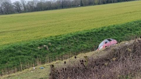 A red car on its side in a ditch between an embankment and a field.