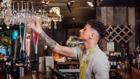 Bar worker reaching up to put glasses on shelf. He has short hair, tattoos on his arm and a yellow apron. The pub has  a wooden interior with soft lighting.