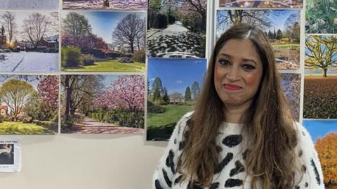 Nadia Gubba with long brown hair wearing a black and white leopard print jumper stands in front of an exhibition of images of nature. She is smiling. 