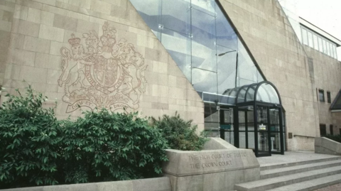 The exterior of a court building. It is grey brick with a crest ingrained into the wall with a glass frontage.