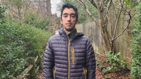 Ali Durrani looking at the camera. He is wearing a Rab outdoor jacket which is dark blue with a yellow zip. He is in a park and there is lots of shrubbery and greenery surrounding him. He is stood near a fence which has black graffiti on it. 
