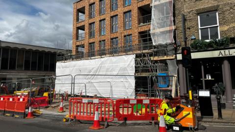 A construction site next to a road. A man in a high-visibility jacket is crouched next to a temporary traffic light pedestrian crossing signal.