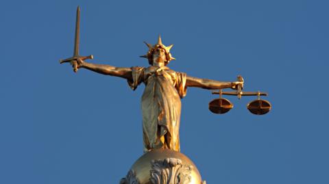 A gold-coloured statue of Lady Justice stands on top of a domed building against a clear blue sky. She is holding a sword in one outstretched hand and a set of scales in the other.