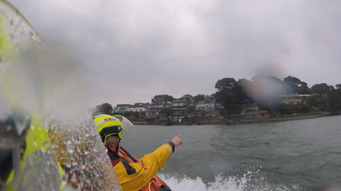 View from a lifeboat crew's camera. Their colleague is pointing towards the shore on a grey, rainy day.