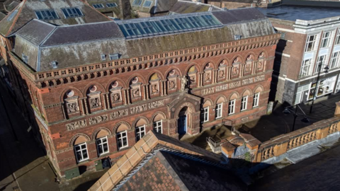 Looking down on a large ornate brick building, with a steeped tiled roof, and a row of large windows.