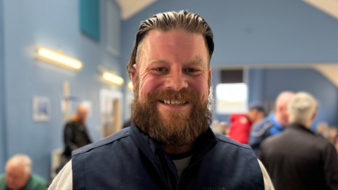 Tommy Barnes, who has slicked back hair and a bushy brown beard, stands in the blue and white painted veterans' centre and smiles. He is wearing a blue gilet on top of a white jumper. In the background are other veterans who are talking with one another.