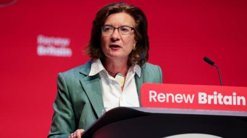 Eluned Morgan wearing a green jacket and a white blouse, in front of a lectern that says "Renew Britain" written in white on a red background.