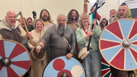 A group of 11 men and women dressed as Vikings, including a man in chain-mail armour and some holding shields and swords, cheer as they stand in a tent. 