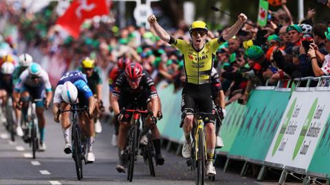 A group of cyclists crossing a finish line. The cyclist in front is wearing a black and yellow outfit. He has his hands in the air and is screaming. Crowds line the barriers on the road.