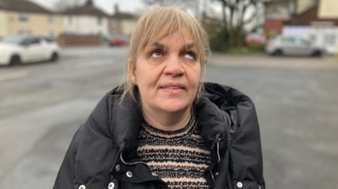 A woman with blonde hair, wearing a black jacket over a stripy style jumper. She is the focus of the picture, with cars and houses blurred in the background.