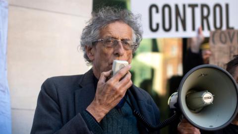 A man in his 70s with white hair and wearing a grey jacket speaks into a megaphone at an outdoor protest. 