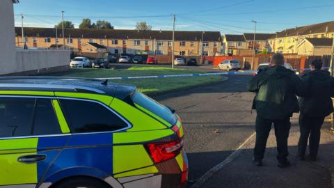 Police car and officers beside police tape in Carnhill housing estate