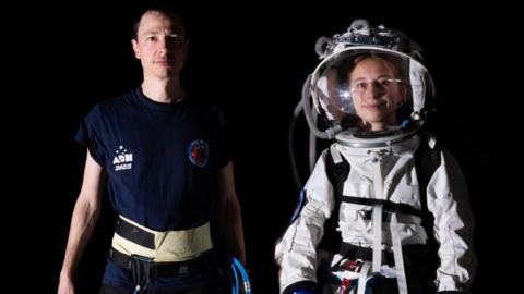A man and women are stood next to each other. One is wearing a white space suit with a glass helmet, the other is wearing a navy blue T-shirt and has piping in his hand. They are both looking directly at the camera and stood in front of a black background. 