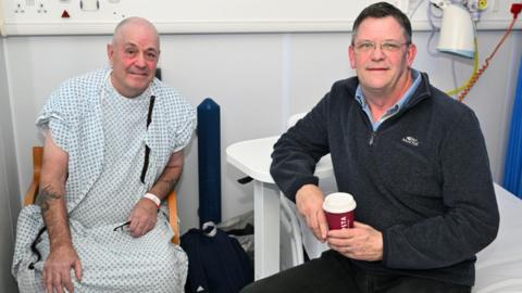 Two men sit side by side in a hospital. One is in a white plastic hospital gown after treatment. 