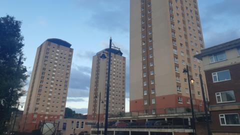Three tower blocks, all of the same design - the lowest few floors of the buildings are coloured orange, while the storeys above are beige. There are multiple windows on each level. A car park is located in front of one of the blocks.