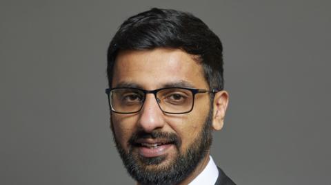 Official portrait of Adnan Hussain taken against a grey backdrop. He has black hair and a beard and is wearing glasses.