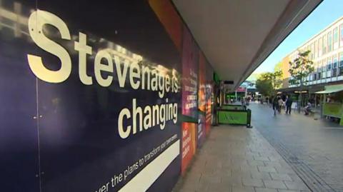 Shopping street in Stevenage. A banner on the wall to the left indicates that "Stevenage is changing". There is a cafe further down the street with green barriers either side of tables and chairs . There is a green food stall to the right in front of three-storey modern shop buildings. Some shoppers can be seen walking on the block-paved pavement.