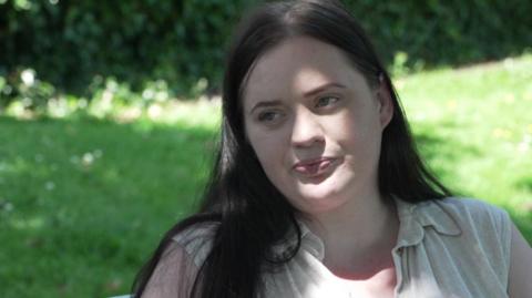 Fiona Goddard, who has long dark hair and is wearing a cream shirt sits on a park bench.