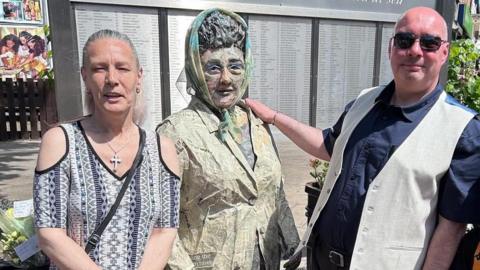 Councillor Tracey Henry, chair of the Headscarf Revolutionary statue management committee, and Ian Cuthbert with a life-sized paper sculpture of Lillian Bilocca. Henry is wearing a purple and white patterned top with cut-out shoulders, a silver cross necklace and a black bag across her body. She has long, grey hair tied back in a ponytail. Cuthbert is bald and is wearing black sunglasses, a navy shirt with the sleeves rolled up and a beige waistcoat undone. 
