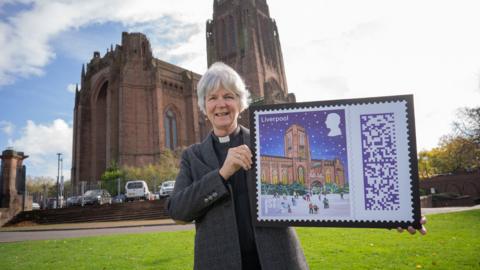 Reverend Dean Sue Jones holds a framed print of the Liverpool Cathedral First Class Royal Mail stamp outside the building.