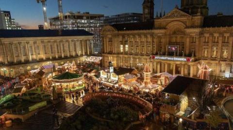 An aerial image of an illuminated Christmas market taken at dusk. Behind the market are Birmingham's town hall and the city's skyline.