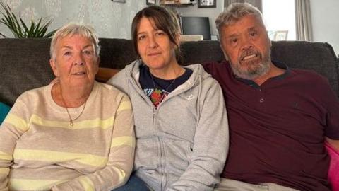 Lin Bruchez sits on a sofa next to her daughter and her husband. Lin has short grey hair and is wearing a lemon and grey striped top. Joanne has brown hair and is wearing a grey top with a black T-shirt and Eddie has a beard and grey hair and is wearing a burgundy top. They are sitting in the living room of their home.