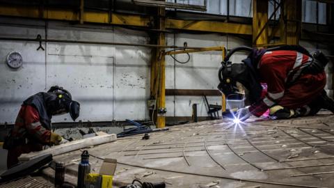 Two workers in boiler suits and protective equipment work on the huge bronze shield. One of them is working with a welding torch and a bright blue spark can be seen in front of him