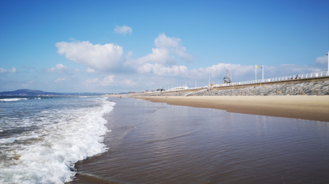 A sandy beach with the sea in front and a stone wall and while fence behind.
