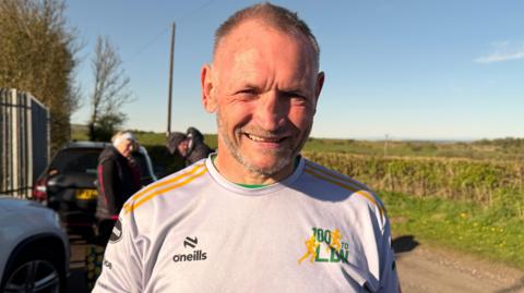 Gary McKee has short grey hair and a beard. He is wearing a grey running top and smiling. Three people are standing next to a parked car on the path behind him.