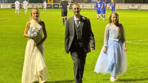 A bride in white dress holding a bouquet of flowers walks off a football pitch. Next to her is her new husband, who is wearing a great suit and tie with a white shirt and next to him is a bridesmaid wearing a light blue dress. In the background are players waiting to kick off.