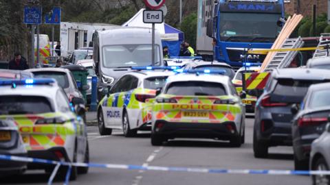 Police cars and other vehicles at the scene on a residential street. A police cordon can be seen at the front.