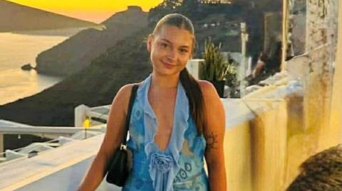 A woman, with dark hair, tied up, wearing a blue dress. There is a cliffside in the background with a sunset visible. 