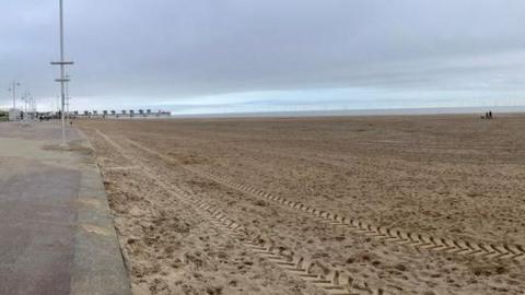 A view of the beach on a cloudy day. A vehicle has left tracks in the sand in the foreground. Part of the promenade is in the left hand side of the picture. The beach is deserted apart from a couple walking their dog in the distance.
