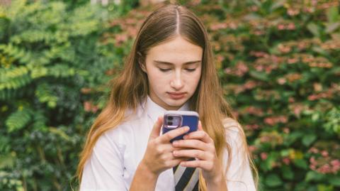A girl in a white school shirt and striped tie with long brown hair holds a phone with a blue phone case. She stands looking at her device with a bush in the background. 