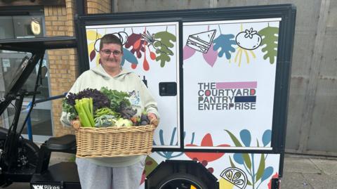 A female volunteer, with glasses and a light green top, holds a basket of vegetables while standing outdoors next to an electric quadricycle, saying THE COURTYARD PANTRY ENTERPRISE on it.