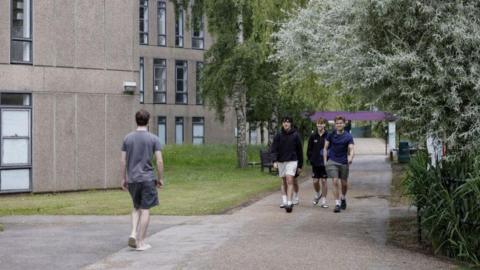 Four male students walk through York University campus. They are wearing shorts and T shirts. The apartment buildings are grey with tall windows.