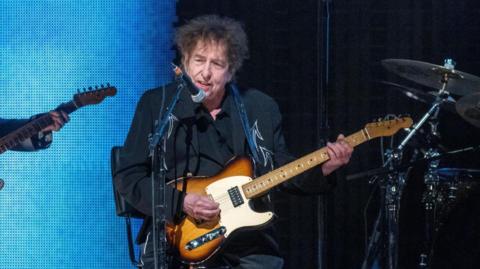 Bob Dylan playing a sunburst guitar while dressed in black and sitting on a stool.