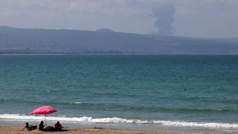 A family sits on a beach in Tyre, Lebanon, as smoke rises on the Lebanese side of the border with Israel during heavy exchanges of fire between Hezbollah and the Israeli military on 25 August 2024