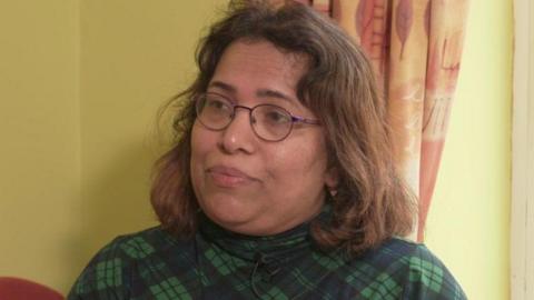 A woman, pictured from the shoulders up, wearing a green and navy top and glasses. She has shoulder length brown hair. There is a yellow wall behind her. 
