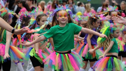 A group of girls wearing green T-shirts and rainbow coloured tutus and make-up dancing in the street during the parade.