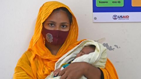 A woman is holding her child in her arms. She is wearing a face covering, and has an orange scarf draped over her head. The child has its eyes closed and is wrapped in a white cloth. 