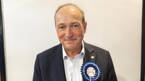 A man wearing a suit and a blue rosette. He is looking towards the camera.