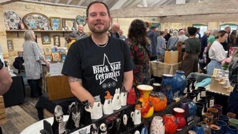 Ian is wearing a black T'shirt with his firm's name and logo on and is stood behind lots of ceramic items on show at the market with crowds in the background