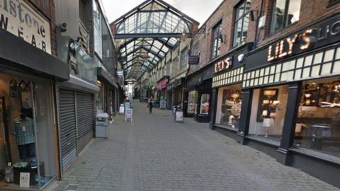 A cobbled street with shops lining each side. There is a glass roof covering some of the buildings. 