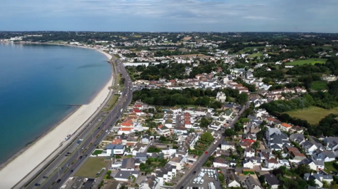 An aerial view of Victoria Avenue - a view of homes and streets along Jersey’s coastline, with a curved beach and coastal road beside the sea. 
