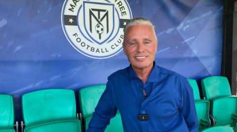 A smiling Barrie Drewitt-Barlow looking straight at the camera. He is sitting on a green seat in a football stadium in front of a blue wall with the logo of Maldon & Tiptree Football Club on it.
