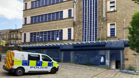 A police van is parked outside an art deco-style derelict department store building in Bradford. Police tape has been used to cordon the building off. 