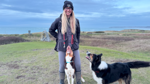 A woman with a dog between her legs and another dog facing her to the right standing on the grasslands and the sea behind her. Grey clouds.