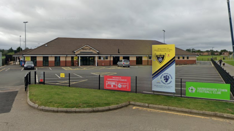 General view of Harborough Town's ground with fencing, parking spaces and club buildings